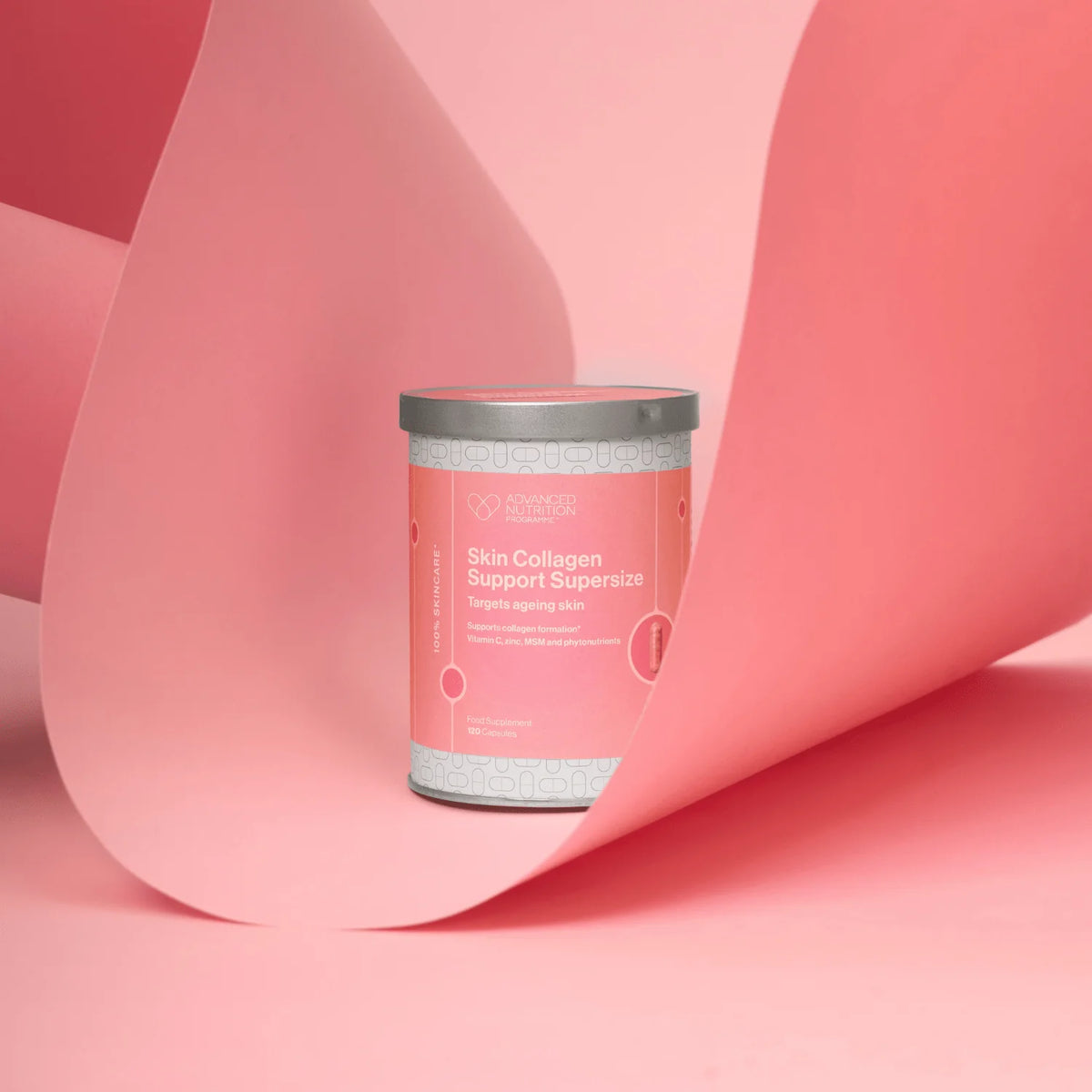 A cylindrical supplement tin labeled 'Skin Collagen Support Supercharge' by the brand 'Vitality', set against a pink studio background with a large pink paper scroll behind it.